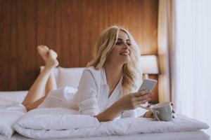 Mulher feliz usando um telefone celular enquanto está deitada na cama e bebe café da manhã foto