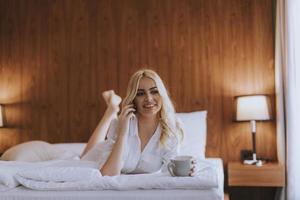 Mulher feliz usando um telefone celular enquanto está deitada na cama e bebe café da manhã