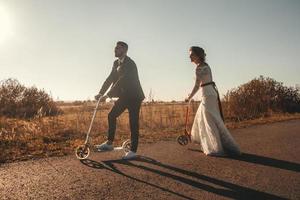 casal de noivos sorridente andando de scooters ao longo da estrada fora da cidade ao pôr do sol. lugar para texto ou publicidade foto