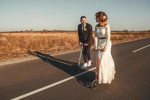 casal de noivos sorridente andando de scooters ao longo da estrada fora da cidade ao pôr do sol. lugar para texto ou publicidade foto