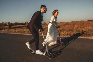 casal de noivos sorridente andando de scooters ao longo da estrada fora da cidade ao pôr do sol. lugar para texto ou publicidade foto