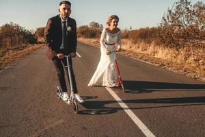 casal de noivos sorridente andando de scooters ao longo da estrada fora da cidade ao pôr do sol. lugar para texto ou publicidade