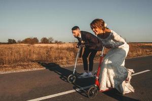 casal de noivos sorridente andando de scooters ao longo da estrada fora da cidade ao pôr do sol. lugar para texto ou publicidade foto