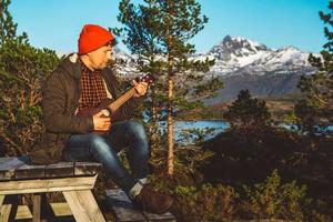 jovem tocando violão, sentado em uma mesa de madeira no contexto de montanhas, florestas e lagos. relaxando e curtindo os dias de sol. lugar para texto ou publicidade foto