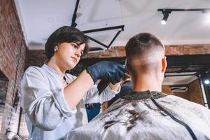 cabeleireira linda corta o cabelo da cabeça da cliente com um aparador elétrico na barbearia. publicidade e conceito de barbearia. lugar para texto ou publicidade foto