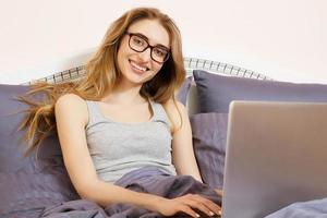 linda garota com óculos deitada na cama com laptop, feliz casual linda mulher trabalhando em um laptop sentado na cama no quarto da casa. mulher trabalhando no laptop. vendas digitais