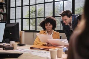 jovem trabalhadora afro-americana senta-se, trabalha com computador, brainstorming, conversando e discutindo com colega caucasiano e parceria sobre empregos de negócios na empresa de escritório do espaço de trabalho. foto