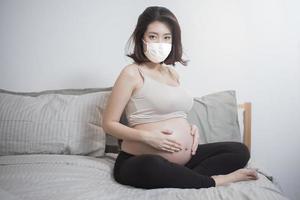 linda mulher grávida asiática está usando máscara protetora em sua casa, conceito de proteção contra coronavírus foto