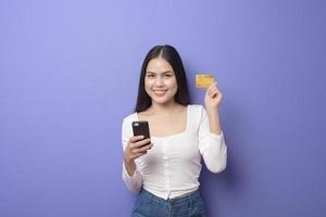 retrato de jovem asiática está usando telefone celular com cartão de crédito em fundo roxo foto