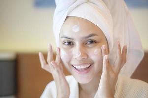 uma mulher bonita feliz em roupão branco aplicando creme hidratante no rosto no quarto, cuidados com a pele e conceito de tratamento