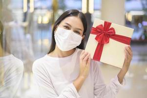 mulher usando máscara protetora segurando uma caixa de presente no shopping, fazendo compras sob o conceito de pandemia covid-19, ação de graças e natal. foto