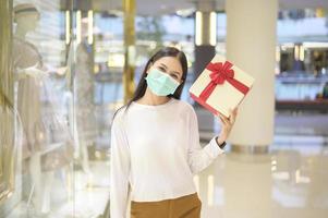 mulher usando máscara protetora segurando uma caixa de presente no shopping, fazendo compras sob o conceito de pandemia covid-19, ação de graças e natal. foto