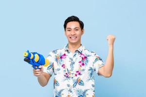 sorrindo bonito homem asiático brincando com pistola de água e levantando o punho isolado no fundo azul do estúdio para o festival songkran na tailândia e sudeste da Ásia foto