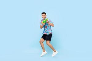 bonito sorridente jovem asiático brincando com pistola d'água e pulando em fundo azul estúdio para festival songkran na tailândia e sudeste da ásia foto
