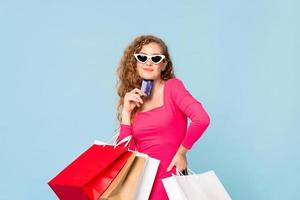 feliz linda mulher caucasiana sorridente com sacolas de compras, mostrando o cartão de crédito na mão isolado no fundo azul foto
