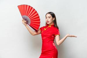 linda mulher asiática de vestido vermelho estilo oriental segurando o ventilador com outra mão aberta para esvaziar o espaço de lado no fundo isolado cinza claro foto