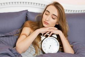 conceito de trabalho dormido demais, insônia de sono ruim - jovem linda garota sonolenta com os olhos fechados segura um relógio e deita-se em sua cama no quarto de manhã foto