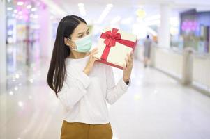 mulher usando máscara protetora segurando uma caixa de presente no shopping, fazendo compras sob o conceito de pandemia covid-19, ação de graças e natal. foto