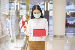 mulher usando máscara protetora segurando uma caixa de presente no shopping, fazendo compras sob o conceito de pandemia covid-19, ação de graças e natal. foto