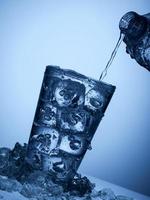 derramando água de uma garrafa de plástico em um copo com gelo em um fundo azul claro foto