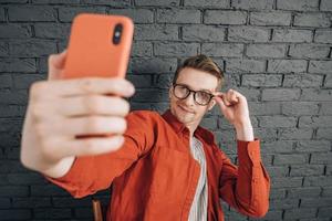 jovem alegre de camisa vermelha e óculos tirando foto de selfie no celular em um fundo de parede de tijolo preto. copiar, espaço vazio para texto