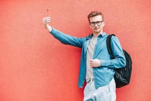 homem de camisa, jeans e óculos carregando uma mochila e olhando para smartphone em pé em um fundo rosa foto