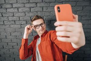 jovem alegre de camisa vermelha e óculos tirando foto de selfie no celular em um fundo de parede de tijolo preto. copiar, espaço vazio para texto