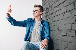 jovem de camisa azul e óculos tirando foto de selfie no celular em um fundo de parede de tijolo preto. copiar, espaço vazio para texto