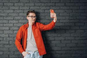 jovem alegre de camisa vermelha e óculos tirando foto de selfie no celular em um fundo de parede de tijolo preto. copiar, espaço vazio para texto