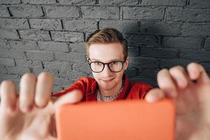 jovem alegre de camisa vermelha e óculos tirando foto de selfie no celular em um fundo de parede de tijolo preto. copiar, espaço vazio para texto