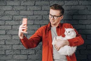jovem alegre de camisa vermelha e óculos segurando um gato fofo branco tirando foto de selfie no celular em um fundo de parede de tijolo preto. copiar, espaço vazio para texto
