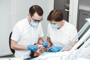 dentista e seu assistente tratam um paciente do sexo masculino em uma clínica odontológica. dentistas em máscaras médicas trabalhando com os dentes do cliente foto