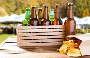 muitas garrafas de cerveja e pacote de batatas fritas na mesa de madeira no fundo de verão turva foto