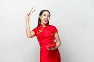 mulher asiática sorridente no tradicional vestido vermelho chinês qipao segurando a caixa de comida de plástico e pauzinhos em estúdio isolado fundo cinza claro foto