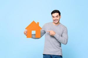 homem caucasiano sorridente segurando e apontando para o modelo de recorte de casa isolado em fundo azul claro foto