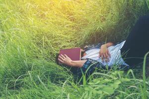 homem jovem hippie deitado na pastagem cochilando cansado depois de ler o livro com a natureza ao redor. foto