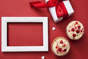 dois cupcakes com creme e decoração, presente, moldura branca simulada em um fundo vermelho. feliz dia dos namorados composição plana foto