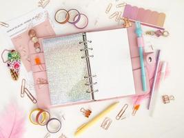 planejador com páginas abertas em um fundo branco e com lindos acessórios canetas, botões, alfinetes e fita colorida. vista superior de um planejador rosa com artigos de papelaria de negócios foto