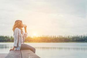 mulher jovem e bonita hipster segurando a xícara de café sentado no lago relaxando com o ar fresco e o nascer do sol da manhã. foto