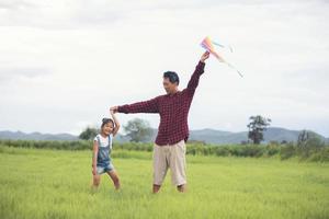 filha asiática e pai com uma pipa correndo e feliz no prado no verão na natureza foto