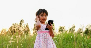 menina criança asiática ouvindo música e cantando de um telefone celular e feliz no prado no verão na natureza foto