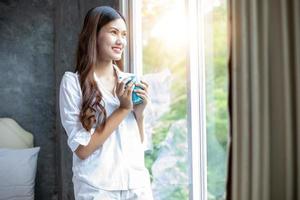 mulheres asiáticas tomando café e acordando em sua cama totalmente descansadas e abrindo as cortinas pela manhã para tomar ar fresco no sol foto