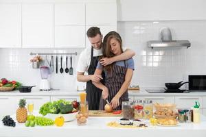 jovem homem caucasiano segurando a namorada dele enquanto ela pica vegetais para preparar o almoço na cozinha. conceito casal junto foto