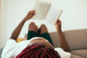 mulher negra deitada no sofá lendo um livro com os pés na parede foto