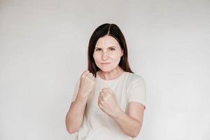 mulher de pé em uma postura de luta, cerrando os punhos com uma expressão agressiva, vestida com camiseta branca casual, posa no interior em um fundo branco foto