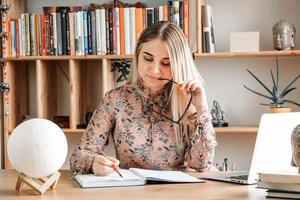 linda mulher vestindo blusa e óculos, sentado à mesa com o laptop e escrevendo algo em seu caderno no fundo das estantes. copiar, espaço vazio para texto foto