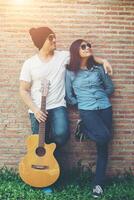 casal hipster está abraçando, olhando para longe e sorrindo em pé ao ar livre tocando guitarra contra a parede de tijolos, namoro passou um ótimo tempo juntos. foto