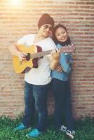 casal hipster está abraçando, olhando para longe e sorrindo em pé ao ar livre tocando guitarra contra a parede de tijolos, namoro passou um ótimo tempo juntos. foto