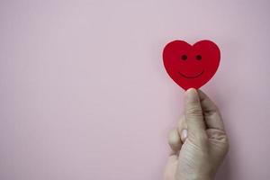 mãos segurando um coração vermelho com ícone de rosto sorridente em fundo rosa pastel. foto