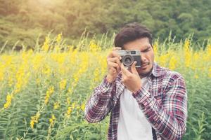 homem jovem hippie bonito fotografando com câmera retro com campo de flores amarelas de natureza. foto